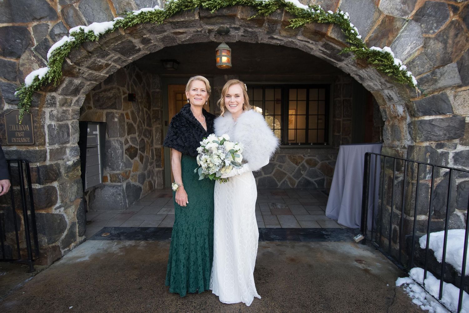 Bride in white gown with fur stole holds bouquet beside woman in green sequin skirt outside stone venue in winter.