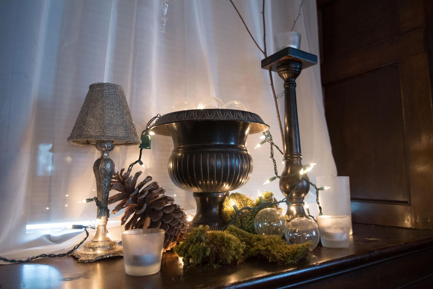 Elegant dark mantle display with brass urn, candlestick, pine cone, moss wreath, and fairy lights on a wooden surface.