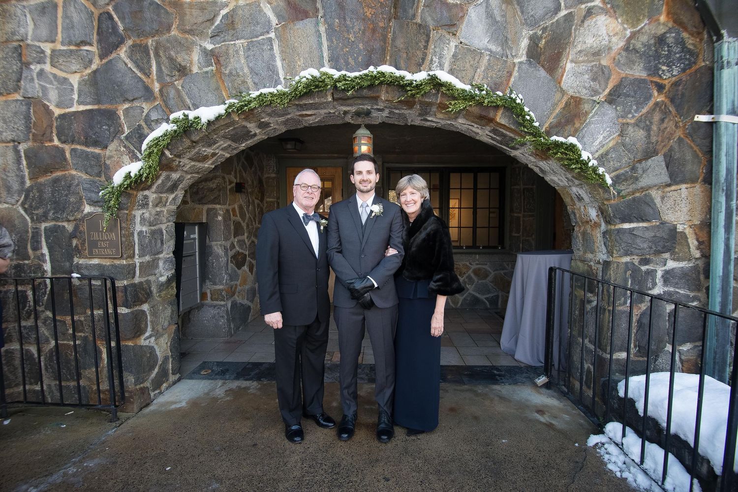 Three formally dressed people posing under a stone arch at a winter wedding venue.