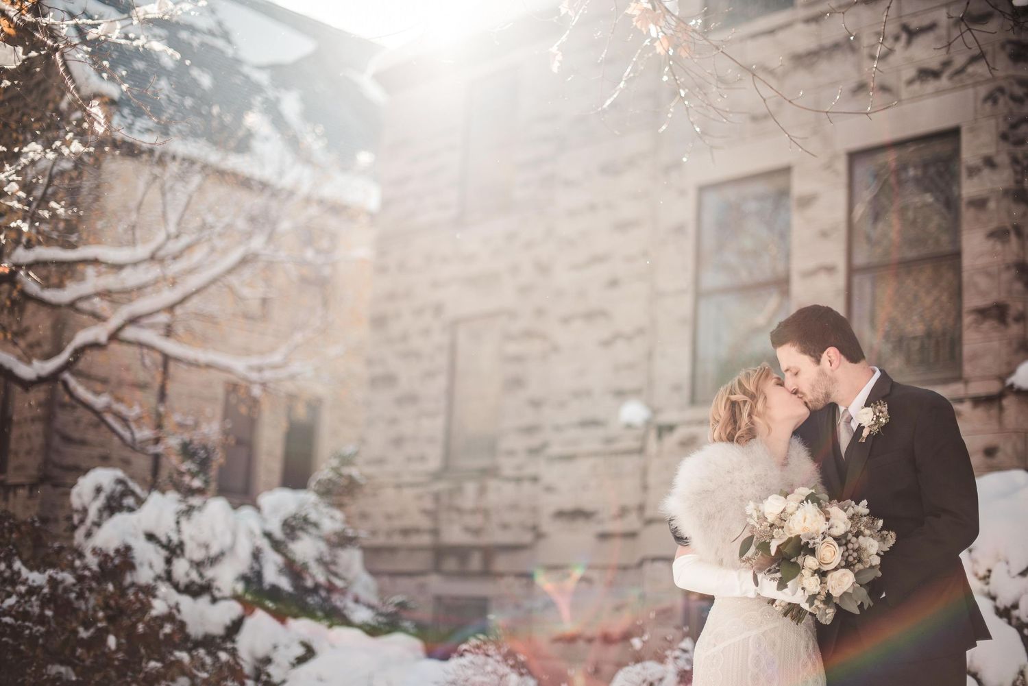 Bride and groom share a romantic kiss in a snowy winter wedding setting outside a stone building.