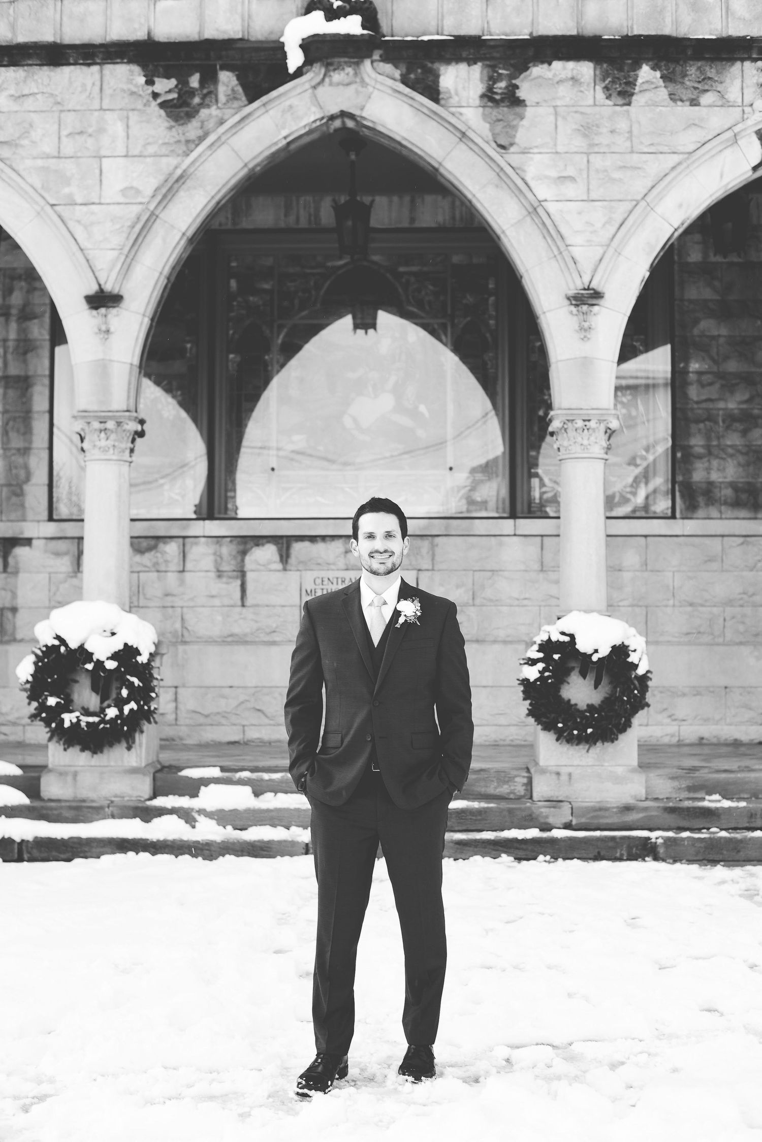 Groom in dark suit stands in snow before Gothic arched building with Christmas wreaths in black and white photo.
