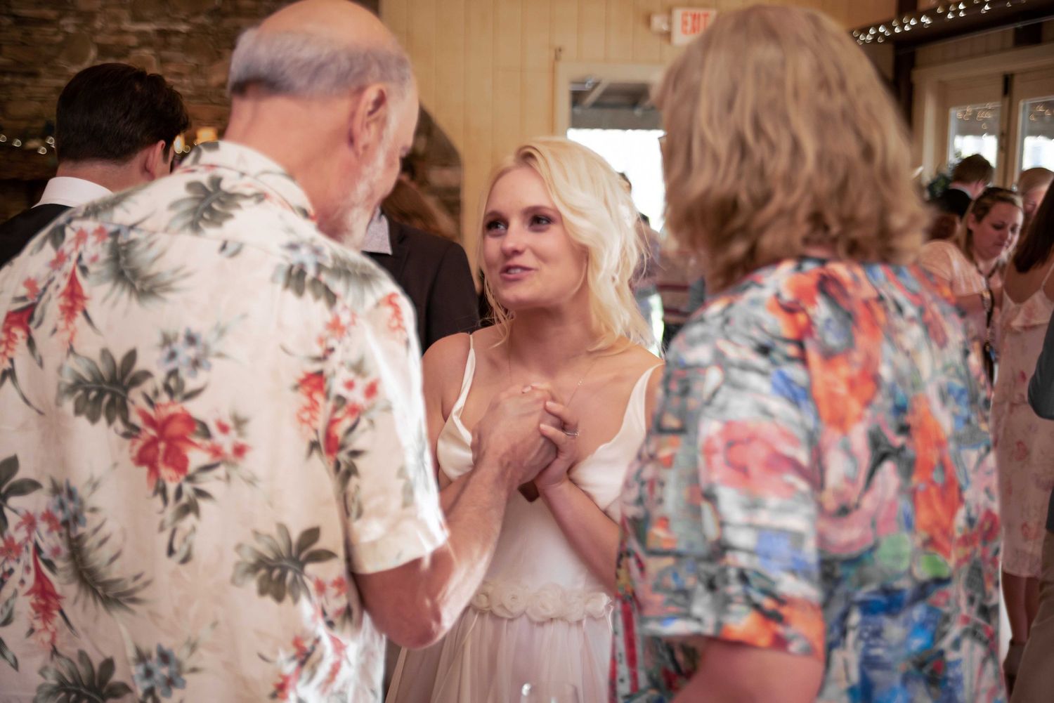 A blonde woman in a white dress converses with guests at an indoor wedding reception.