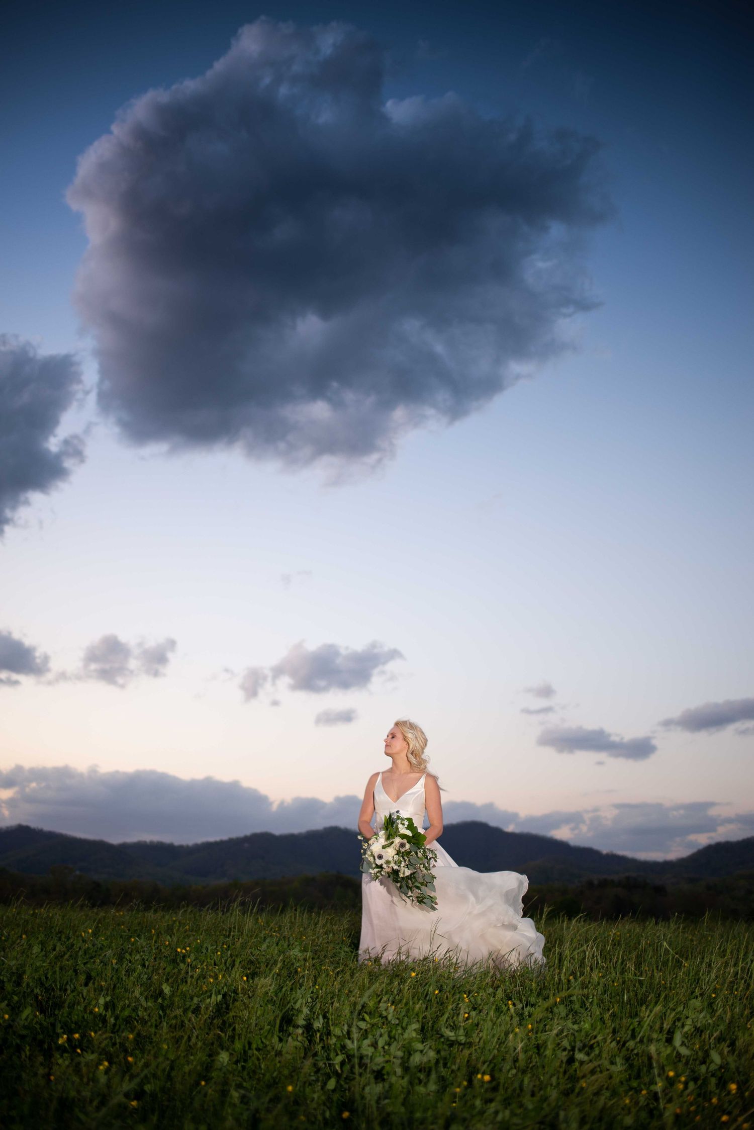 Bride in flowing white gown holds bouquet in green field at dusk, dramatic storm cloud above mountain backdrop.