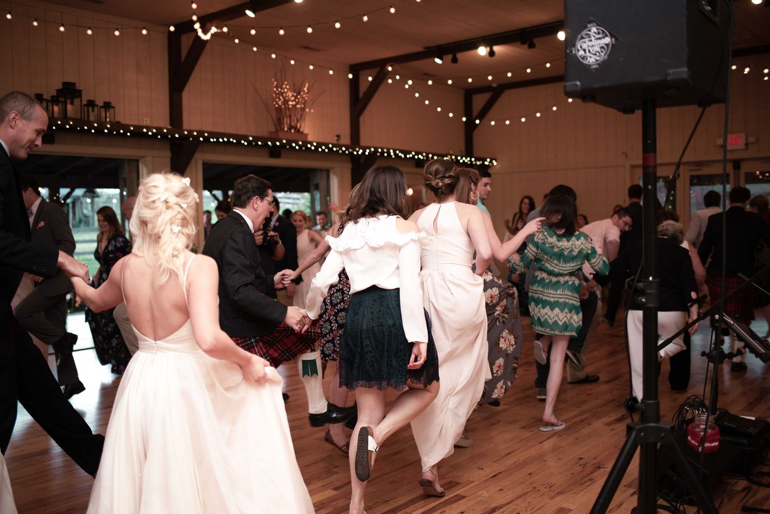 Wedding guests dance joyfully in a rustic barn venue decorated with string lights during a lively reception.