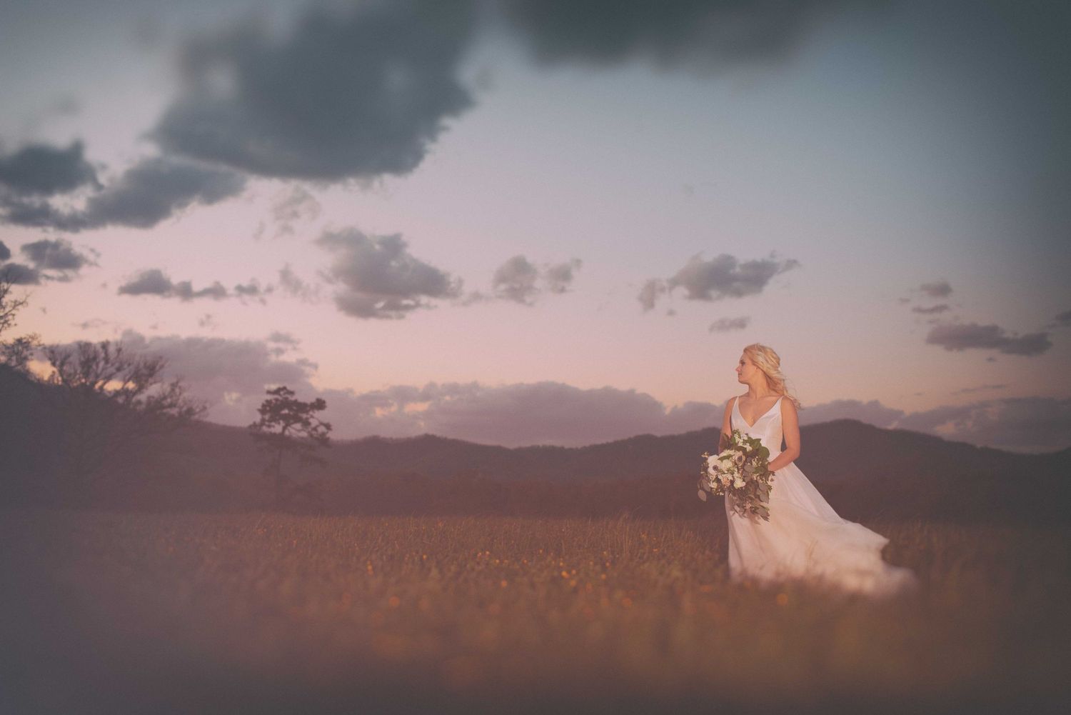 Bride in white gown holding bouquet stands in golden field at sunset with misty mountains in background.
