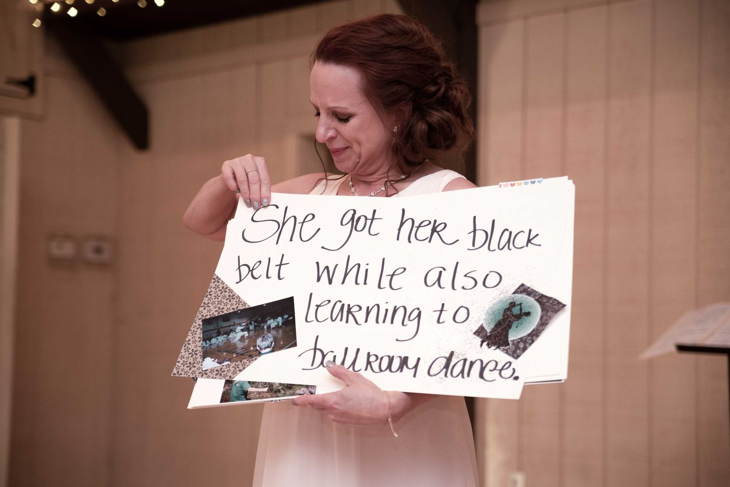 Woman holding a handwritten sign reading she got her black belt while also learning to ballroom dance at an indoor event.