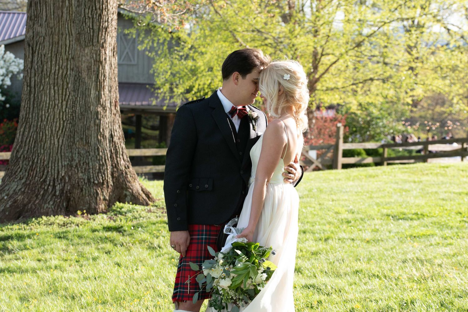 Bride and groom sharing a kiss outdoors, groom wearing a red tartan kilt, bride holding a green bouquet.