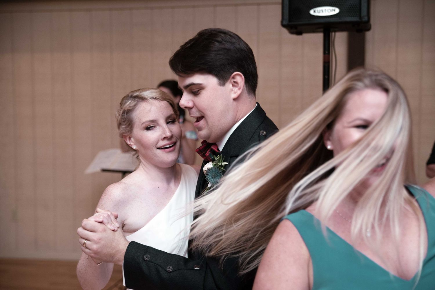 Bride and groom share a dance together at their wedding reception while a blonde woman in teal dress stands nearby.