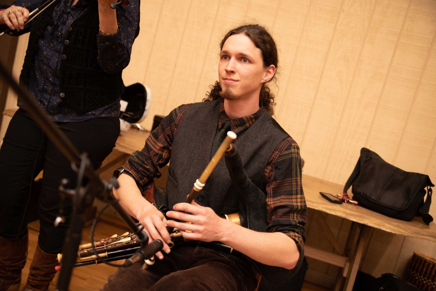 Man with long hair playing a wooden flute instrument while seated, wearing a dark vest over a plaid shirt.