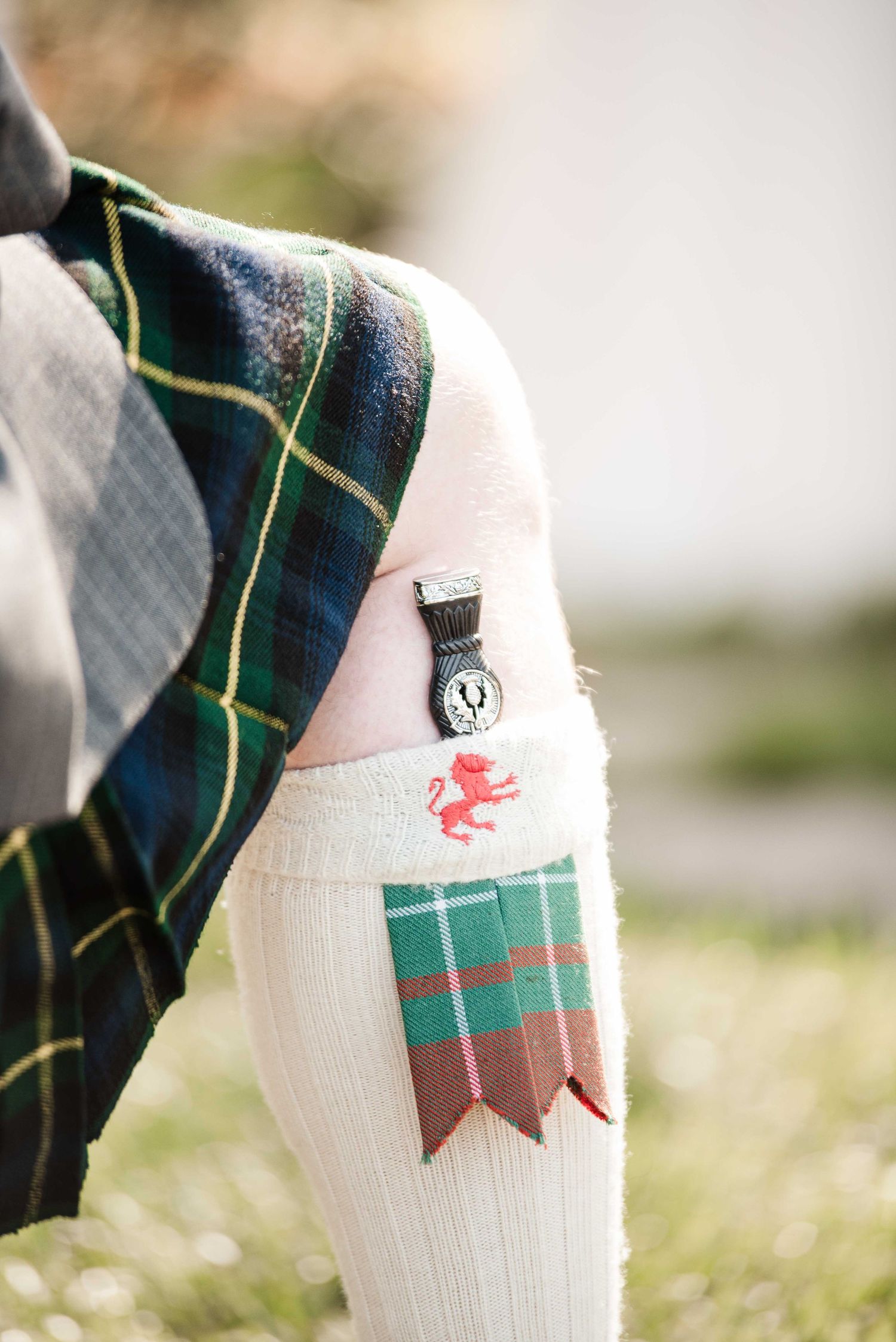 Close-up of Scottish kilt sock with red lion emblem, decorative garter flash, and clan badge pin on knee.