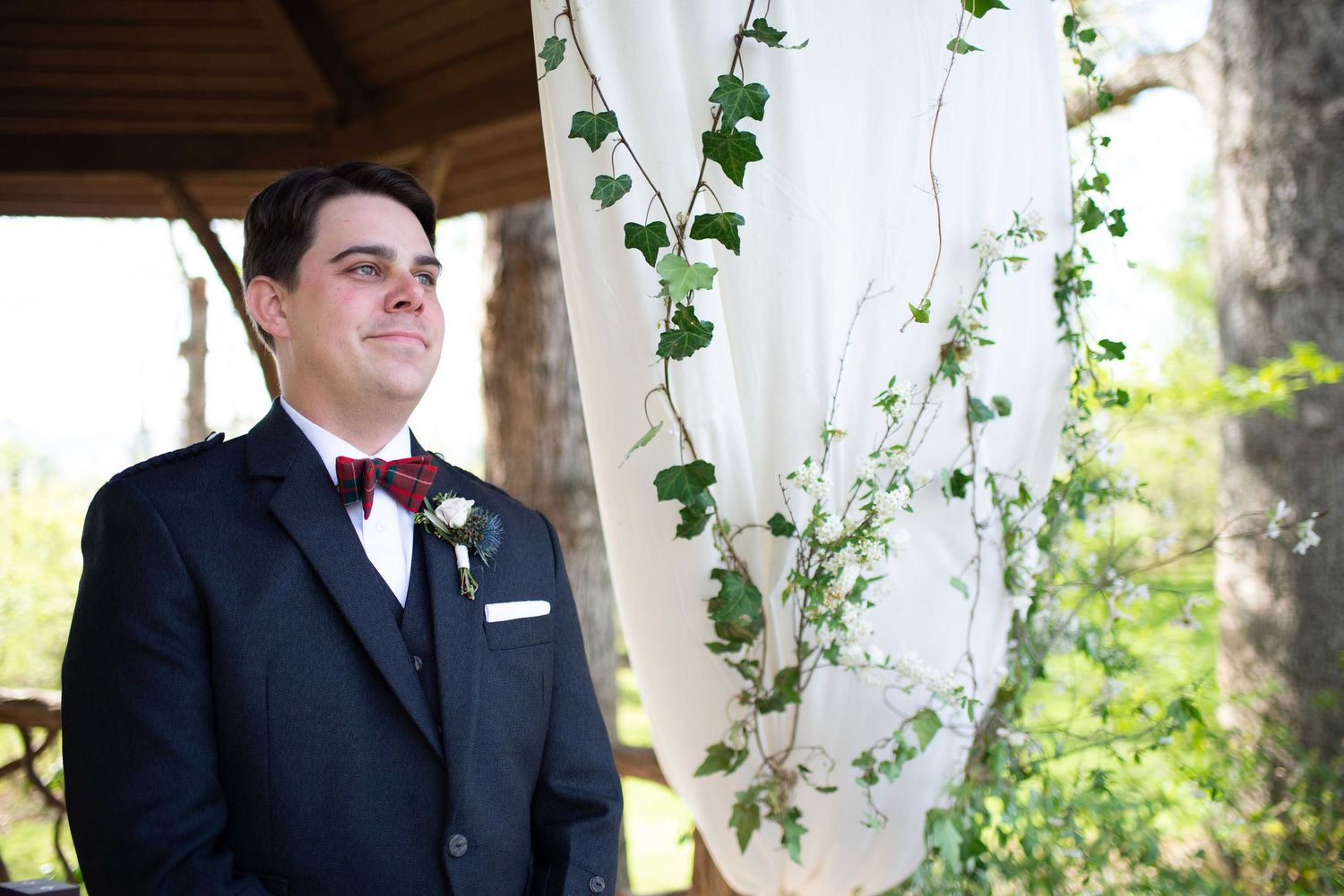 Groom in black suit and burgundy bow tie standing at outdoor wedding ceremony altar decorated with ivy and white draping.