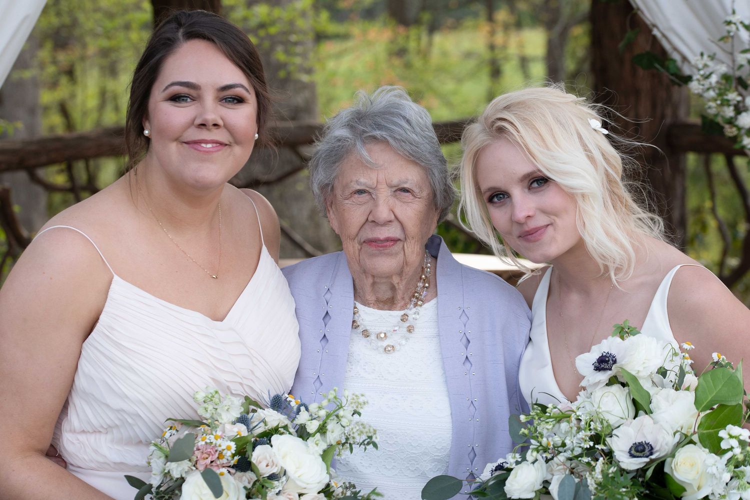 Two bridesmaids in white dresses holding floral bouquets pose with an elderly woman at an outdoor wedding ceremony.
