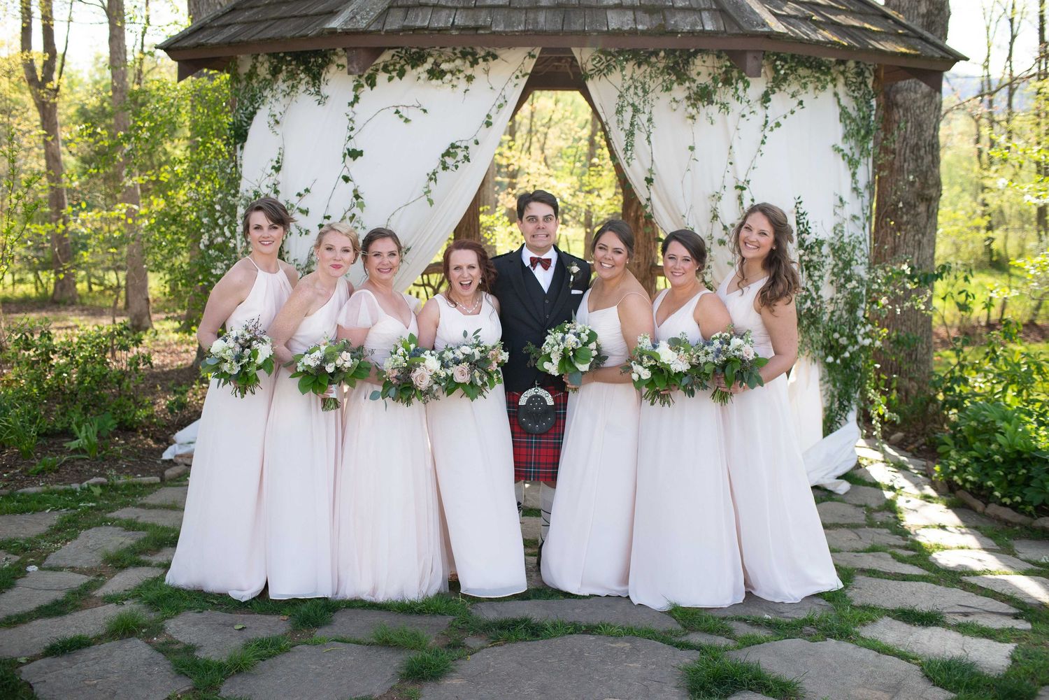 Groom in kilt stands with six bridesmaids in blush gowns holding bouquets at an outdoor wedding gazebo.