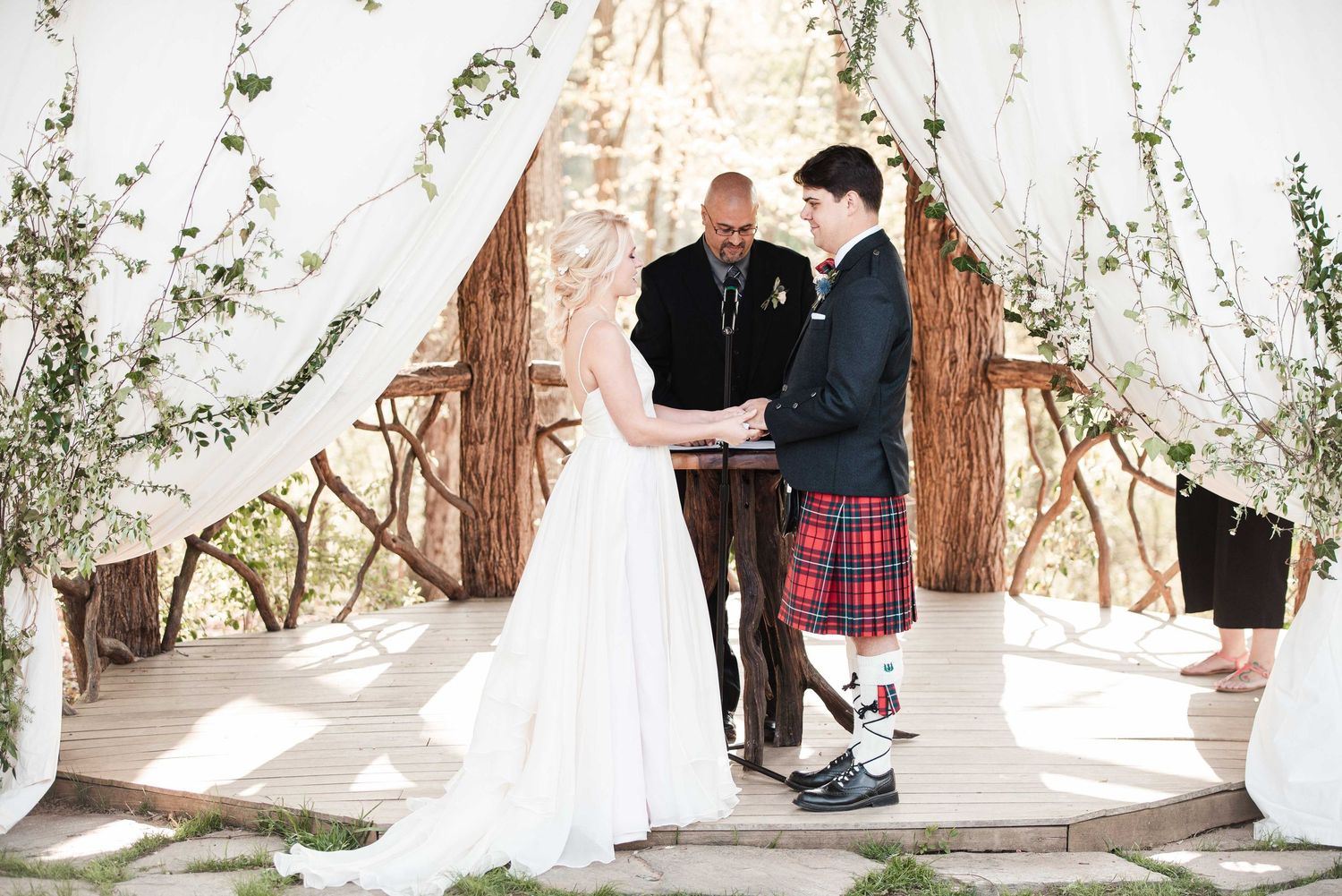 Bride and groom exchange vows under rustic floral arch, groom wearing traditional red tartan kilt at outdoor wedding ceremony.