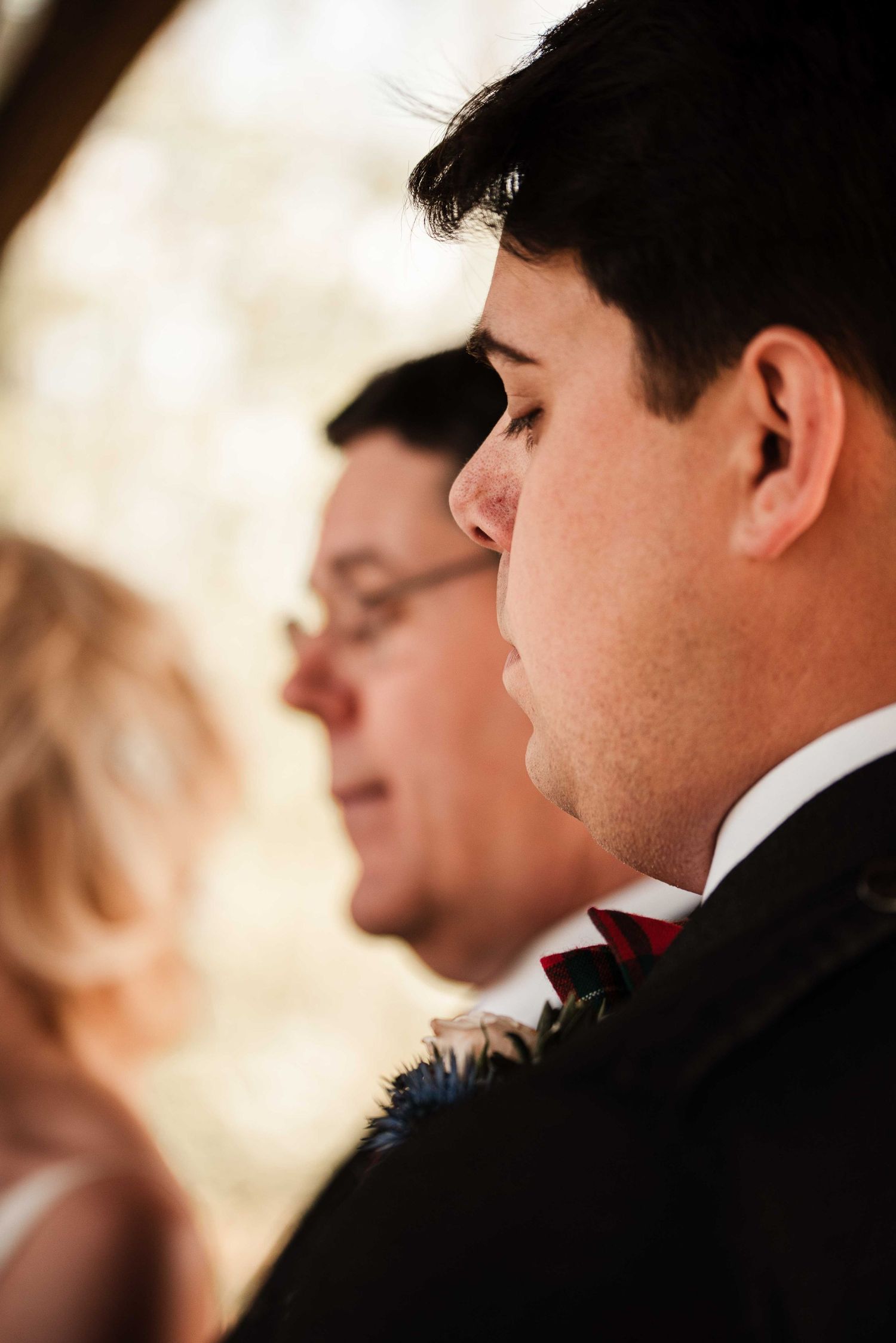 Groom in black suit and red bow tie stands in profile during outdoor wedding ceremony with guests behind.