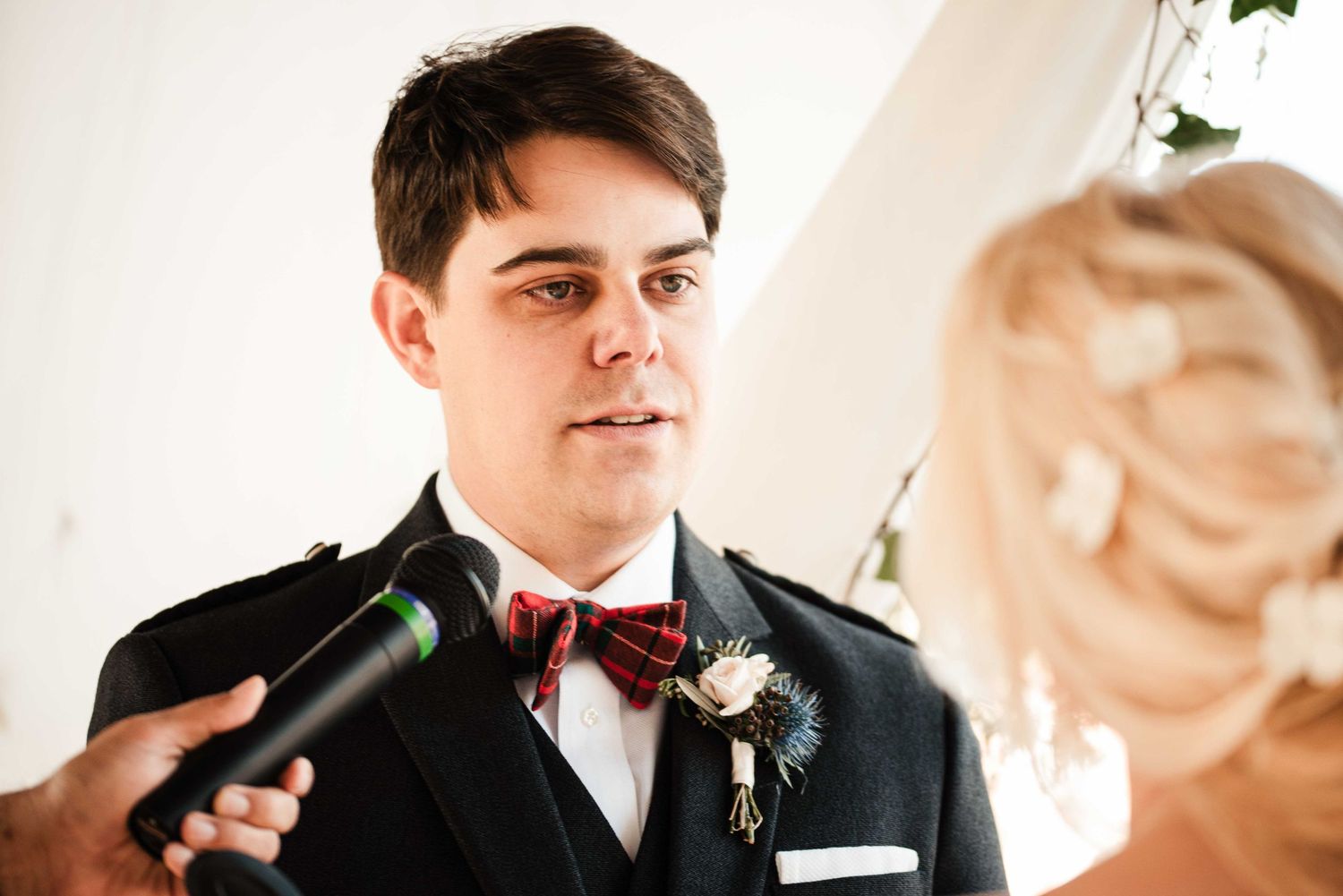 Groom in black suit with red bow tie and boutonniere listening attentively during wedding ceremony vows.