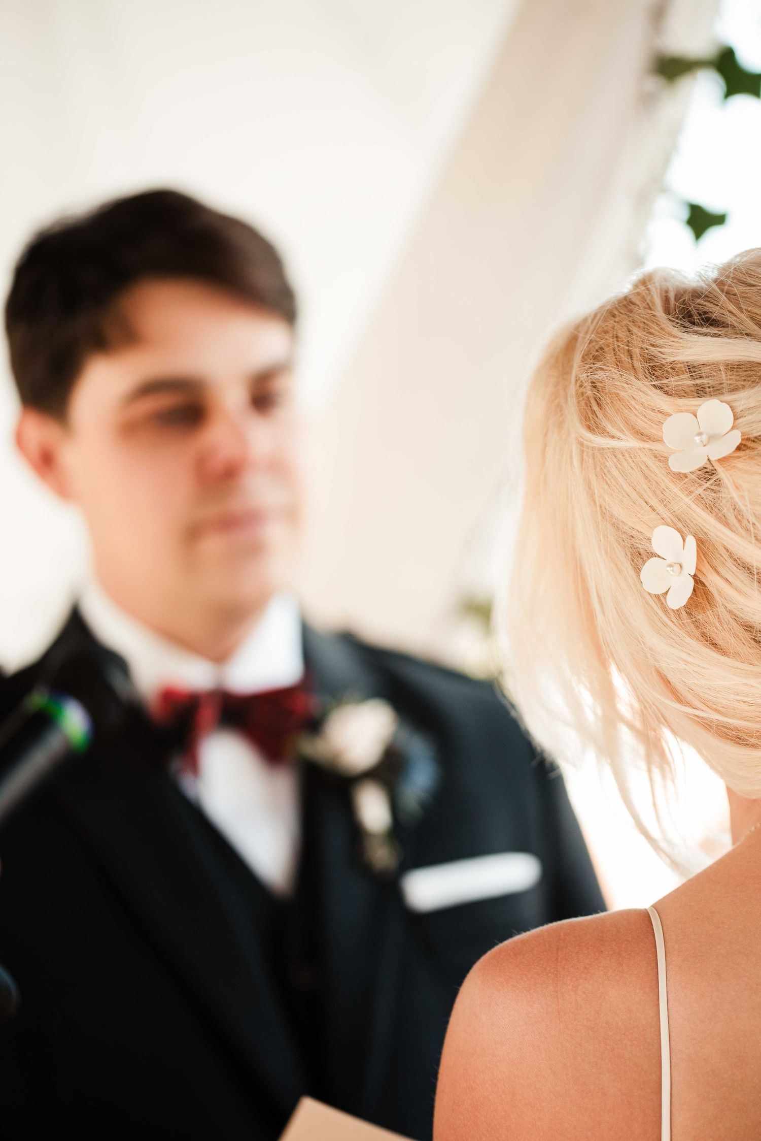 Bride with floral hair pins faces groom in black tuxedo and red bow tie during wedding ceremony.