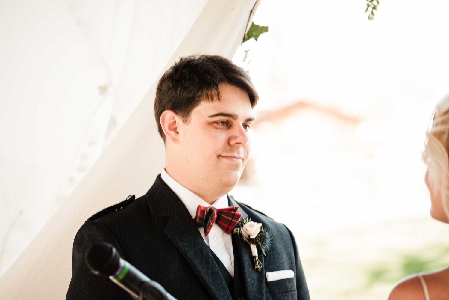 Groom in black jacket and burgundy bow tie with floral boutonniere smiling during outdoor wedding ceremony.