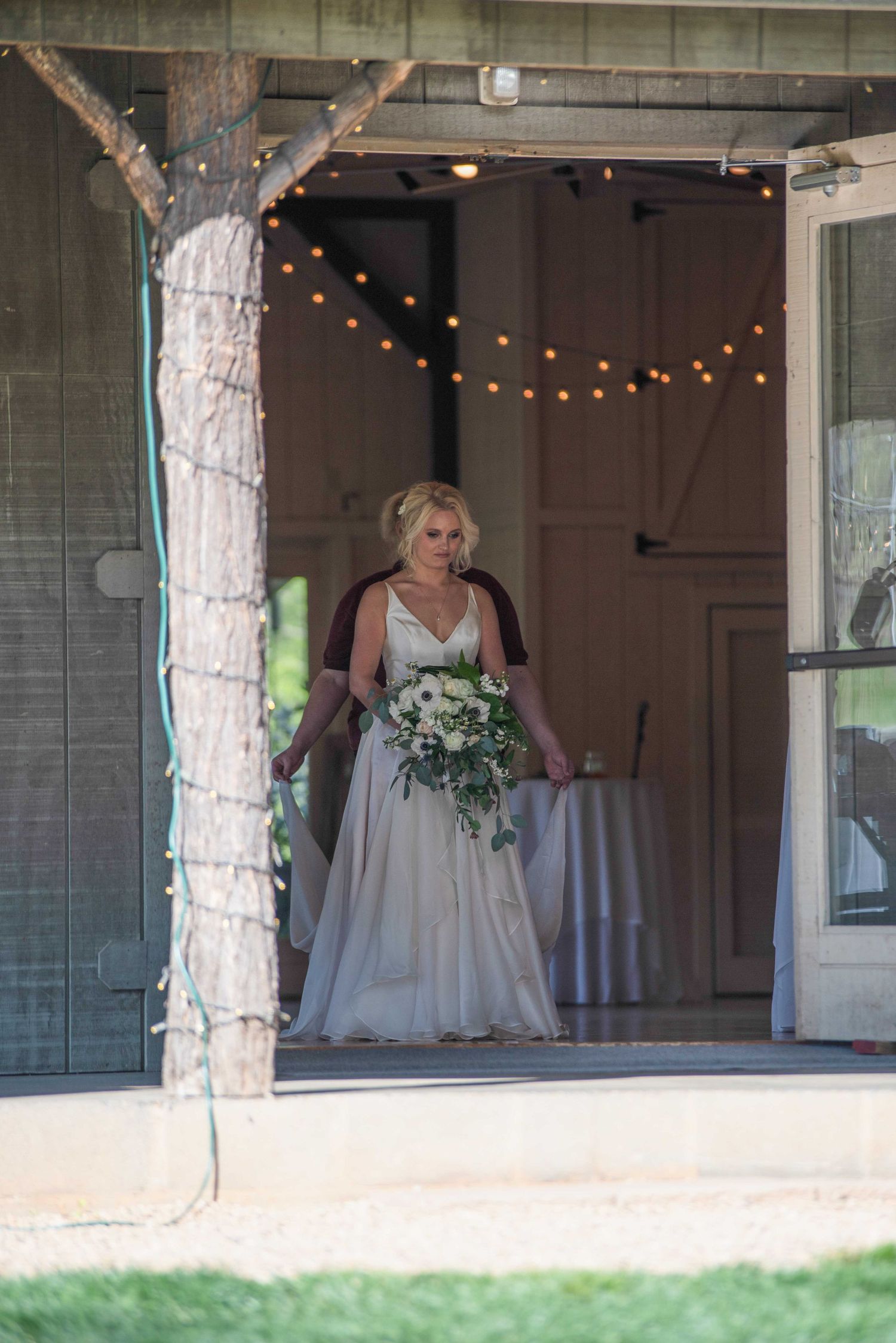 Bride in white gown holding floral bouquet standing in barn doorway with string lights at rustic wedding venue.