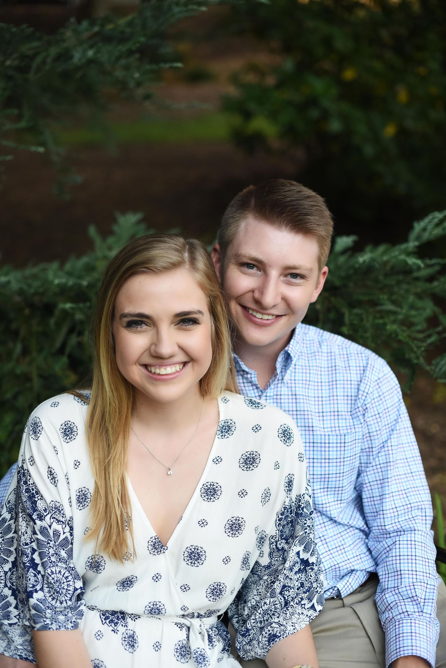 Smiling young couple posing together outdoors surrounded by lush green evergreen shrubs.