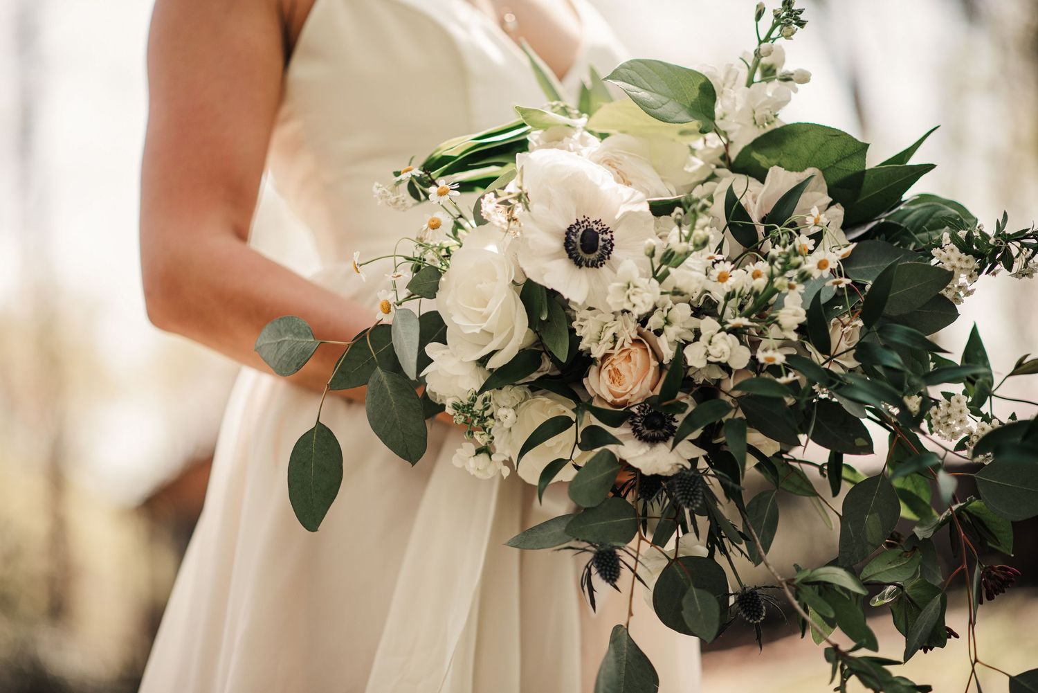 Bride holding a lush bridal bouquet of white anemones, roses, and greenery in a white wedding gown.