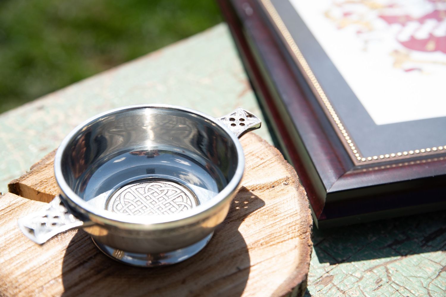 Silver quaich cup on wooden slice beside framed artwork at Scottish-themed outdoor wedding ceremony.