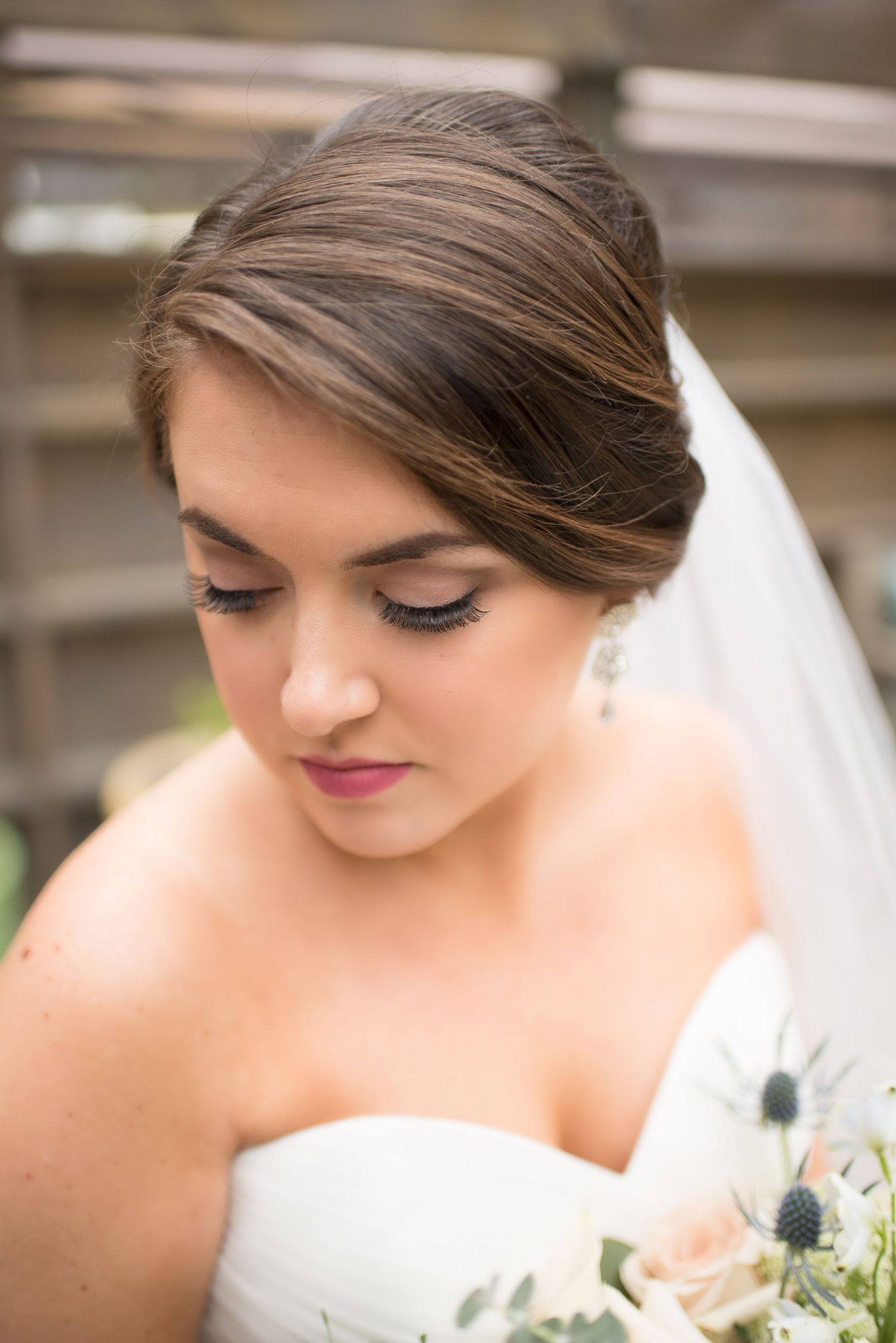 Close-up portrait of a bride with elegant updo hairstyle, veil, and chandelier earrings holding a bouquet.