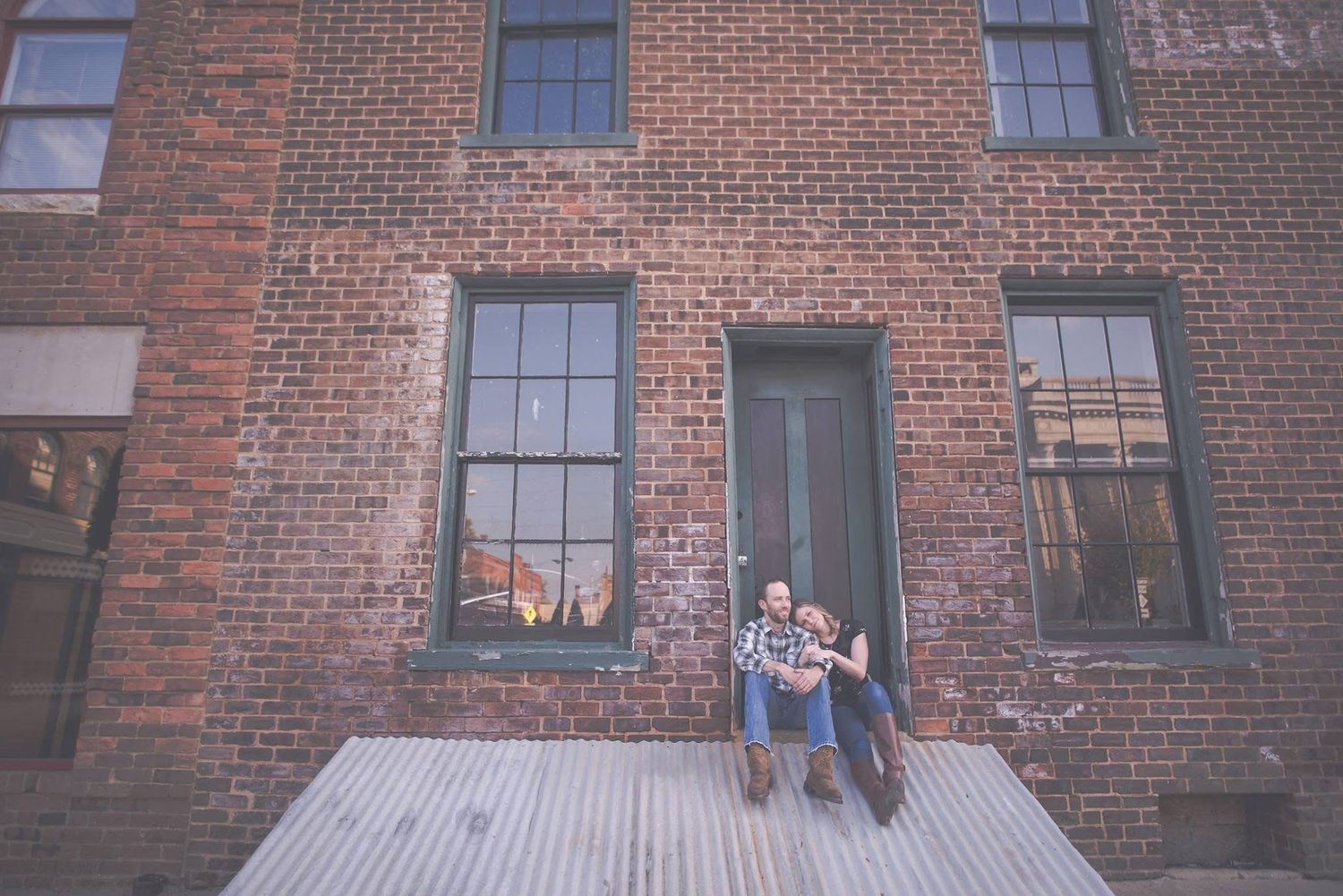 Couple sits together on steps of a rustic red brick building during engagement session.