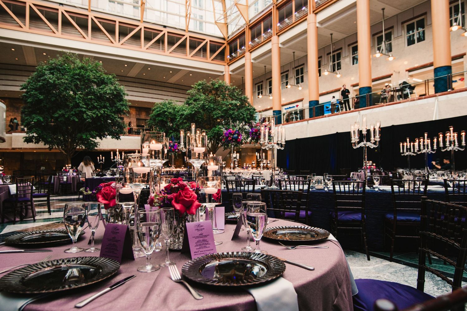 Elegant wedding reception in grand atrium with purple linens, black chairs, and floral centerpieces.
