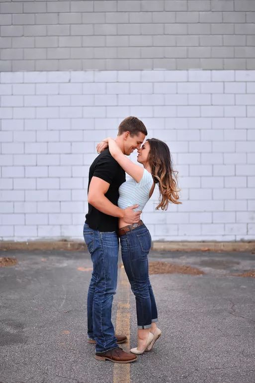 Couple nearly kissing while embracing in front of a white brick wall outdoors.