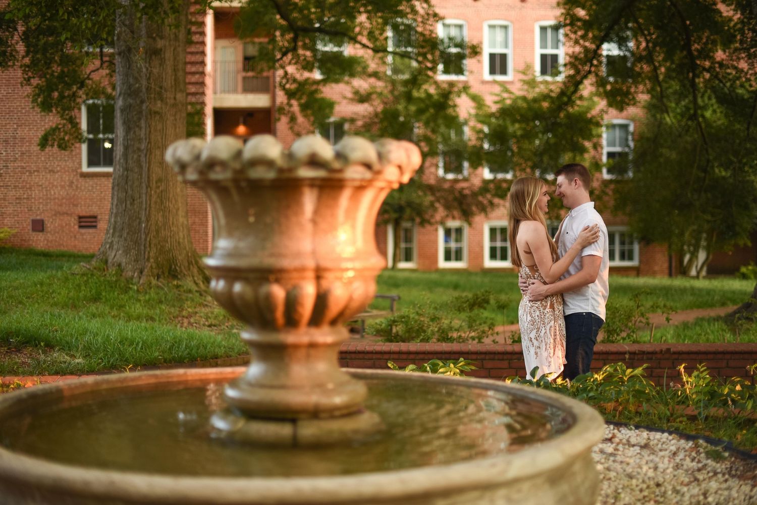 Couple embracing near a decorative stone fountain at golden hour on a brick building campus.