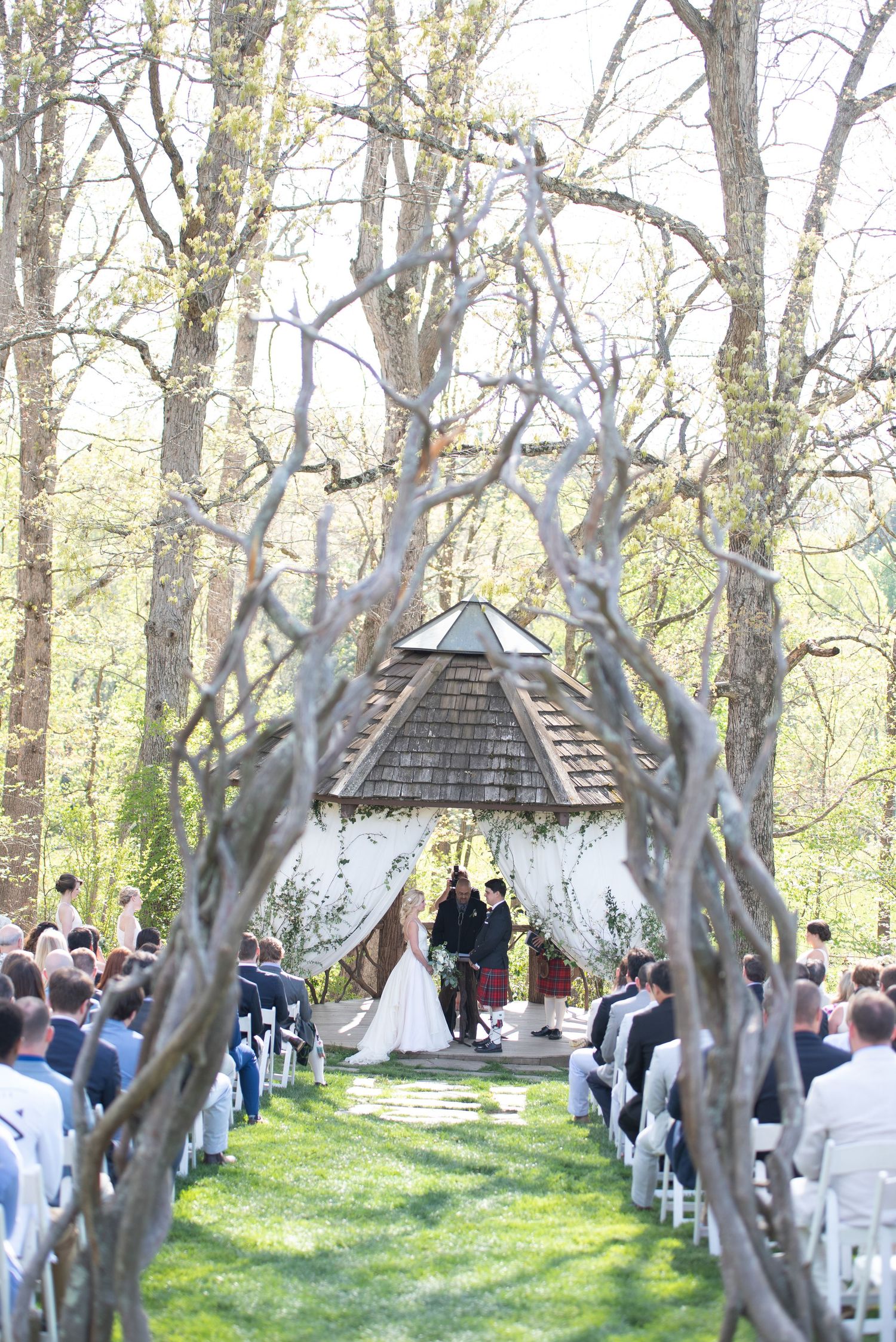 Scenic outdoor wedding ceremony in woodland setting with guests seated along aisle leading to rustic gazebo.