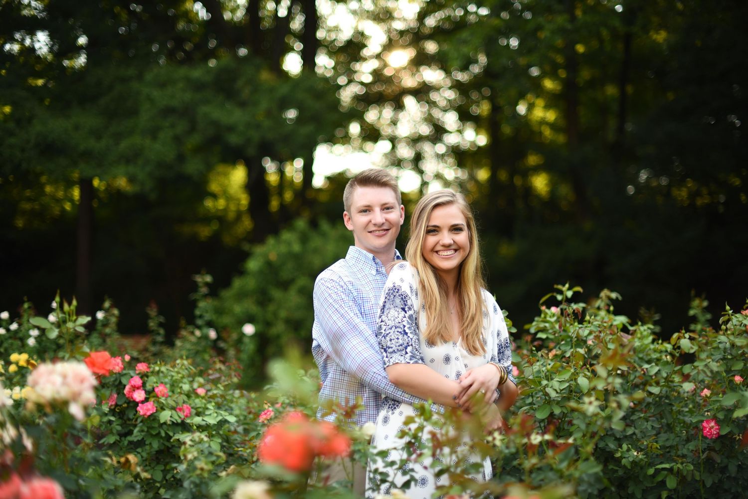 A smiling couple poses together in a lush rose garden with colorful blooms and green trees in the background.