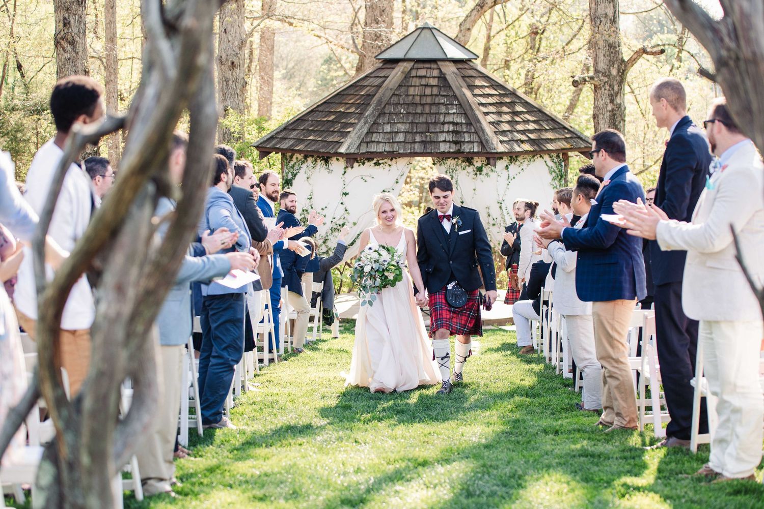 Bride and groom walk down outdoor aisle under gazebo surrounded by cheering guests at a spring garden wedding ceremony.