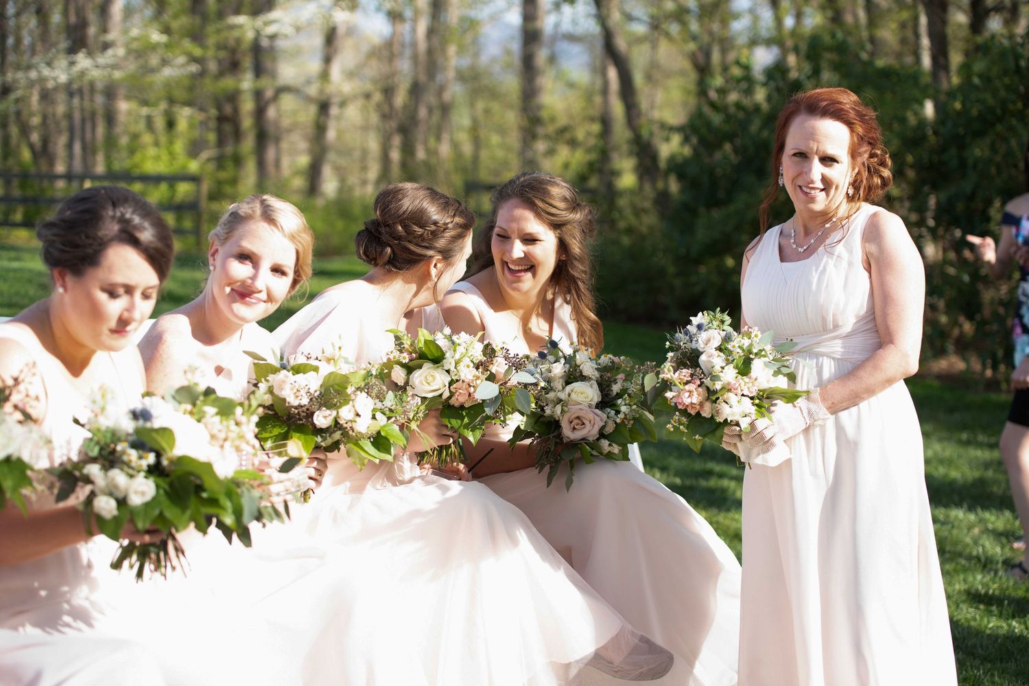 Bridesmaids in blush gowns laugh together holding floral bouquets at an outdoor wedding celebration.