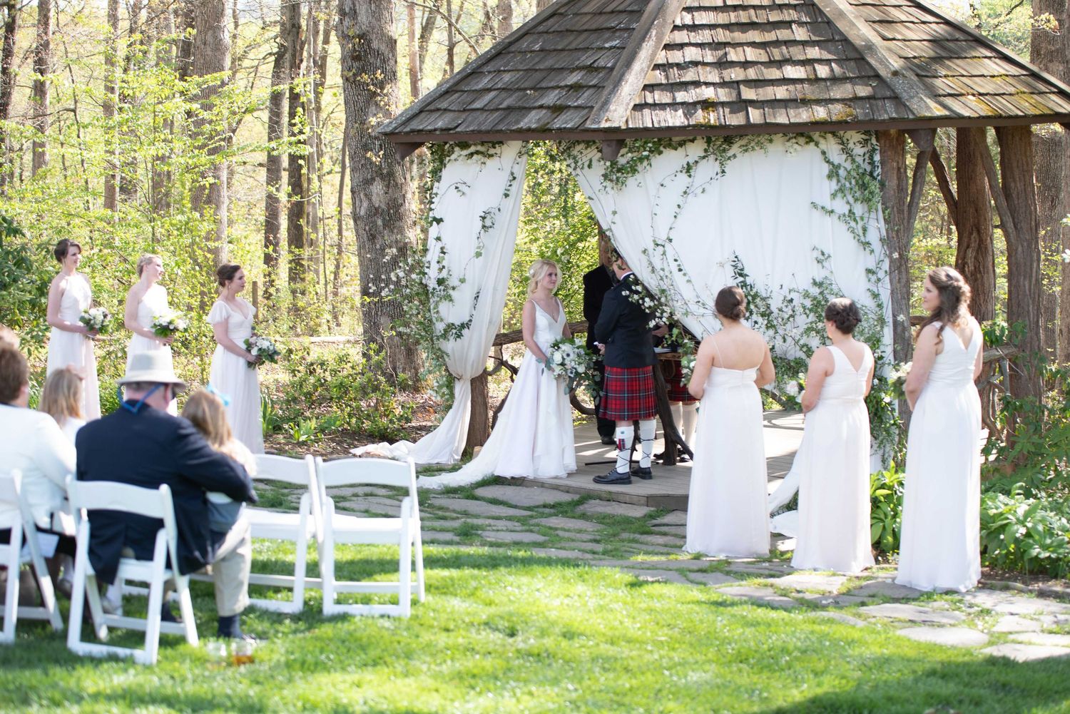 Outdoor wedding ceremony at a woodland gazebo with bridesmaids in blush gowns and guests seated on white chairs.