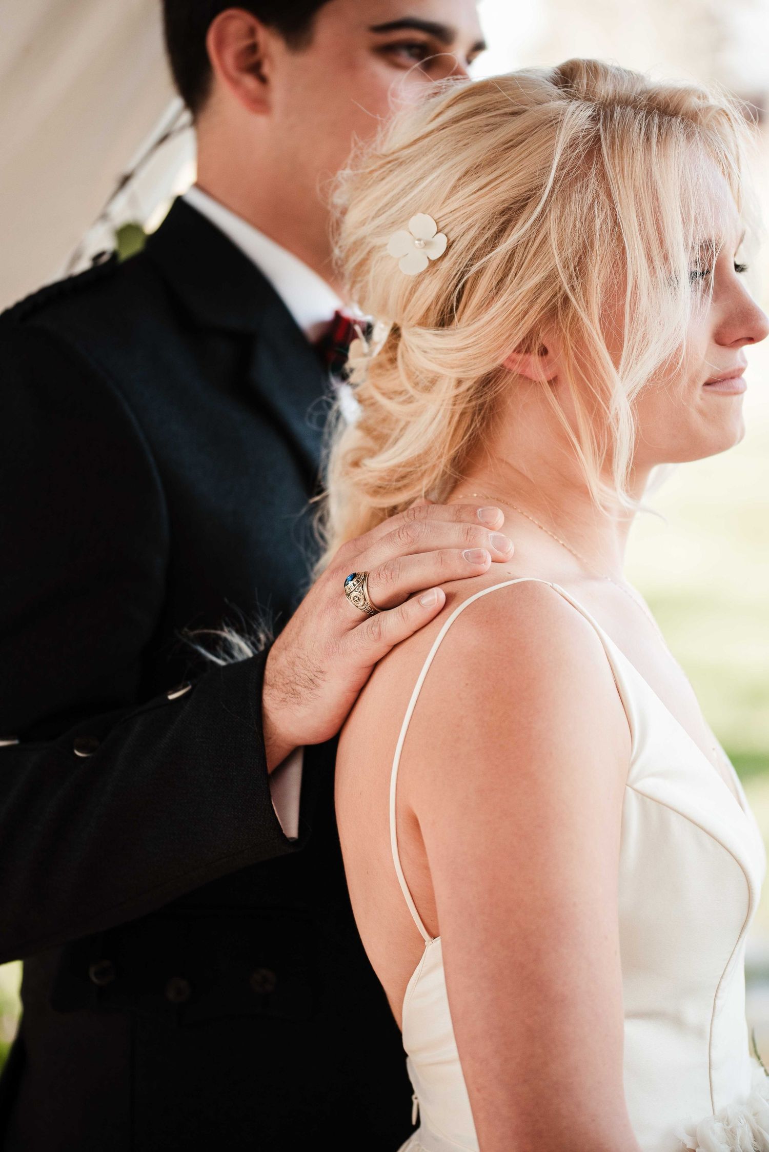 Bride and groom in elegant wedding attire, groom placing hand on bride's shoulder, showcasing a stunning ring.