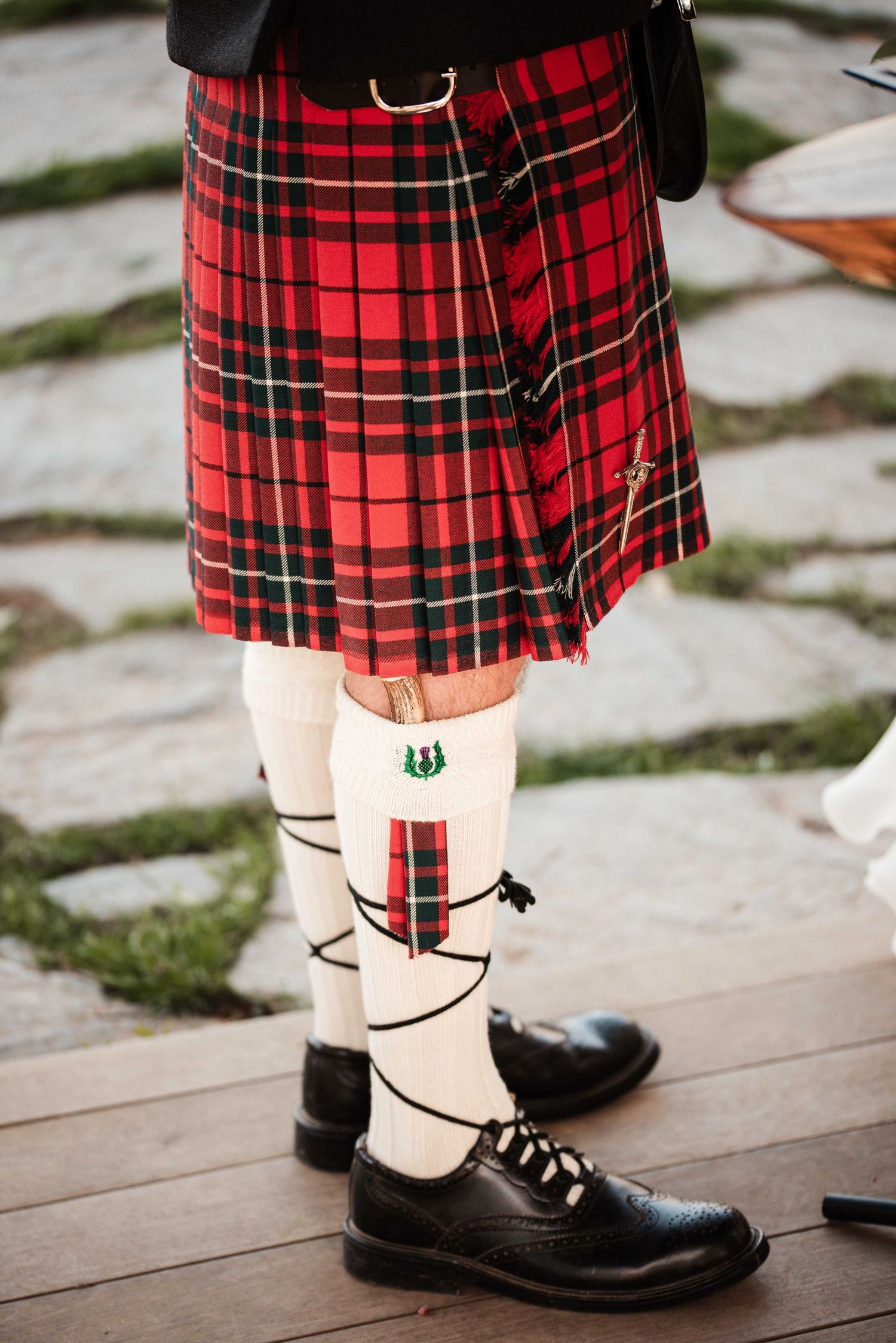 Man wearing traditional Scottish red tartan kilt with white knee socks and black ghillie brogues on stone pathway.
