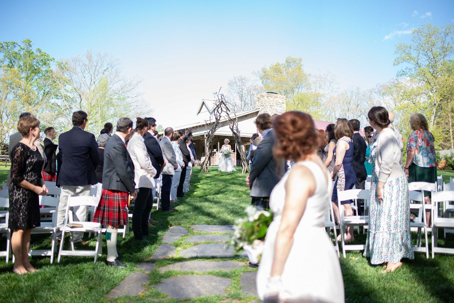 Outdoor wedding ceremony with guests seated in white chairs along a garden aisle on a sunny day.