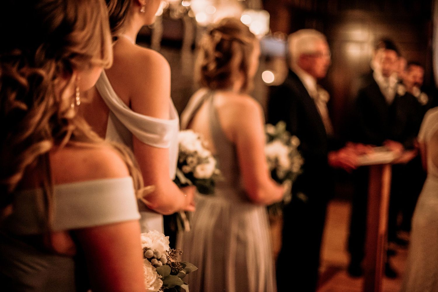 Bridesmaids in elegant gowns holding white bouquets stand at a wedding ceremony aisle.