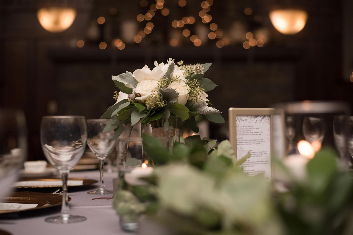 Elegant wedding reception table with white floral centerpiece, crystal glassware, and soft candlelight in a romantic venue.