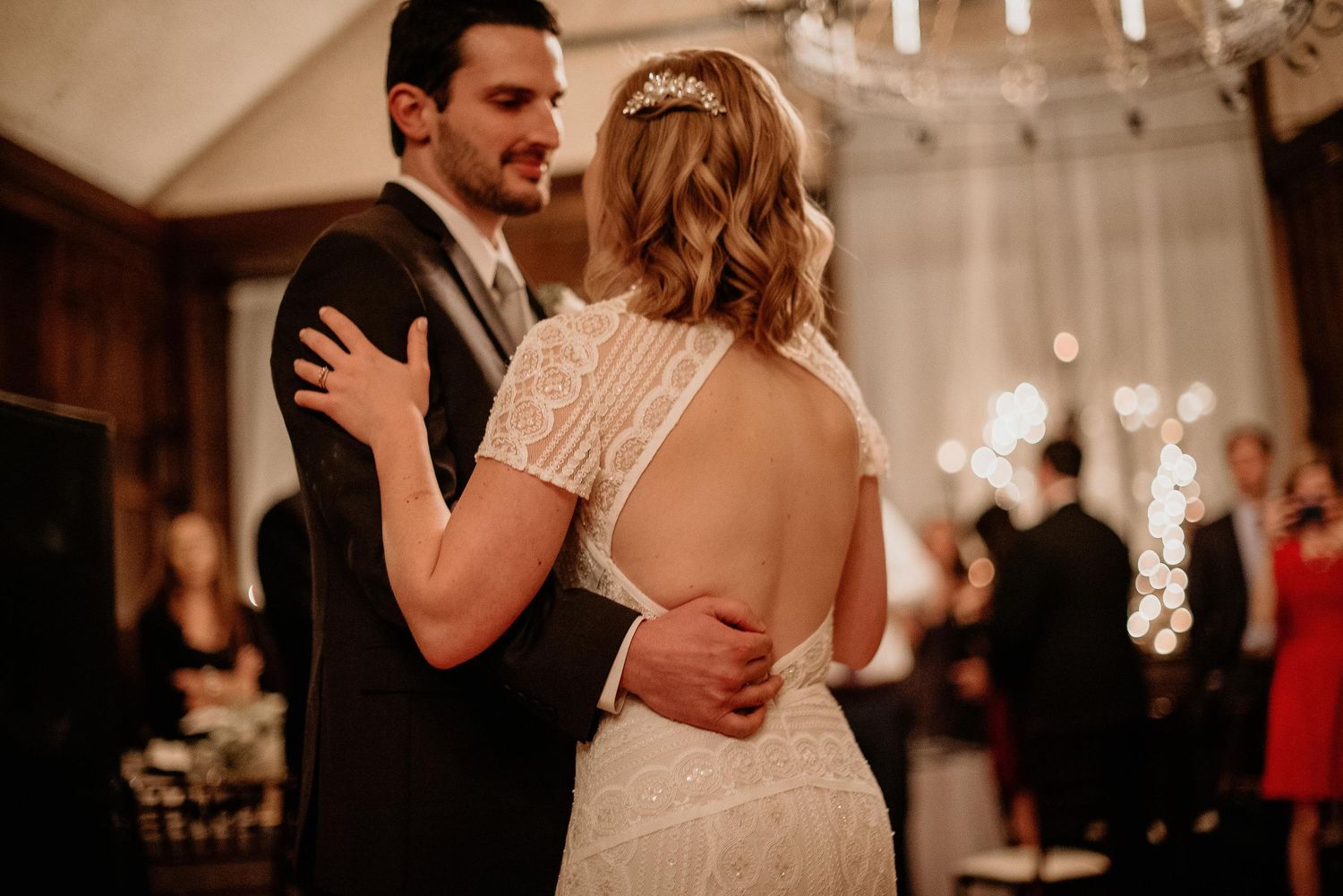 Bride and groom share first dance at elegant wedding reception, bride wearing backless lace gown with hair accessory.