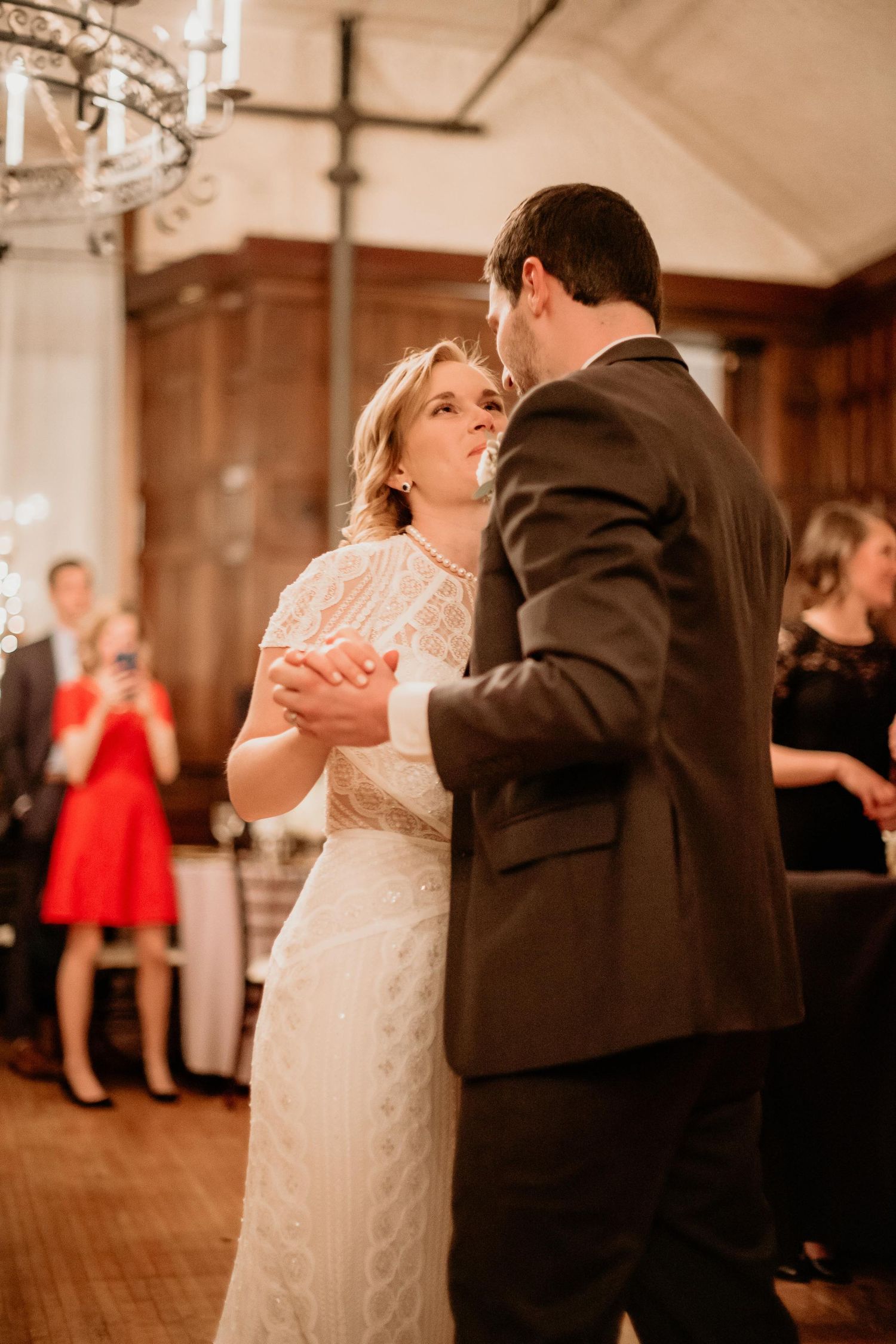Bride and groom share their first dance at wedding reception in elegant wood-paneled ballroom with guests watching.