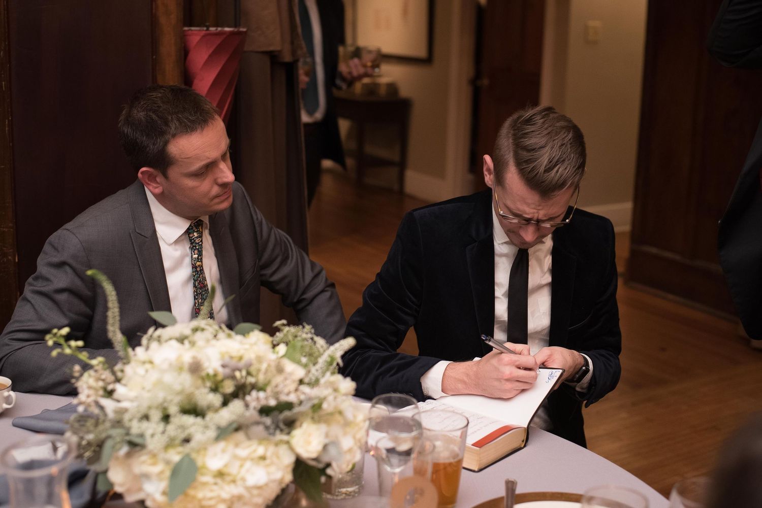 Two men in suits sit at a table, one signing a book at a formal event or wedding reception.