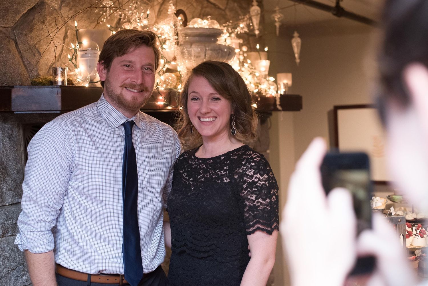 A smiling couple poses together at an indoor event while someone photographs them with a smartphone.
