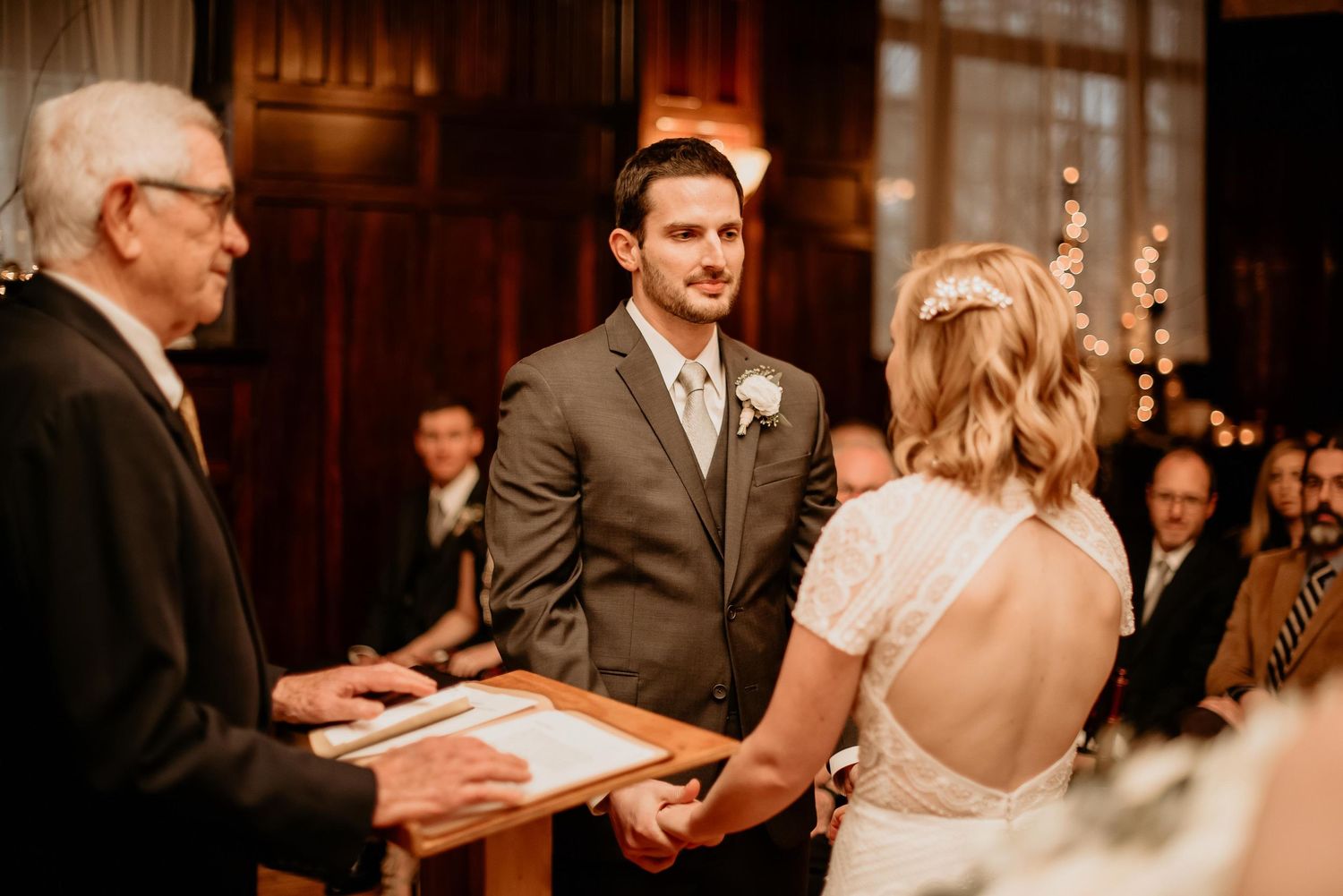 Groom in gray suit holds hands with bride at wedding altar while officiant reads ceremony vows.