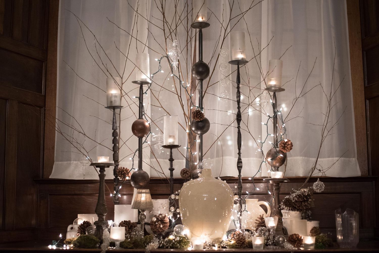 Elegant winter candle display with tall candlesticks, glowing tea lights, ornaments, and bare branches with fairy lights.