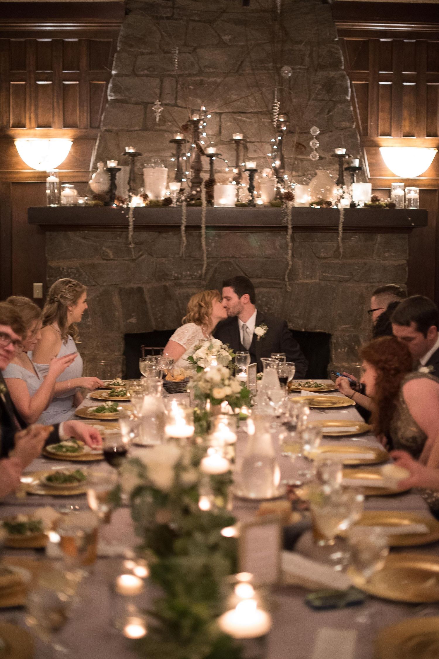 Bride and groom share a kiss at candlelit wedding reception dinner table with stone fireplace backdrop.