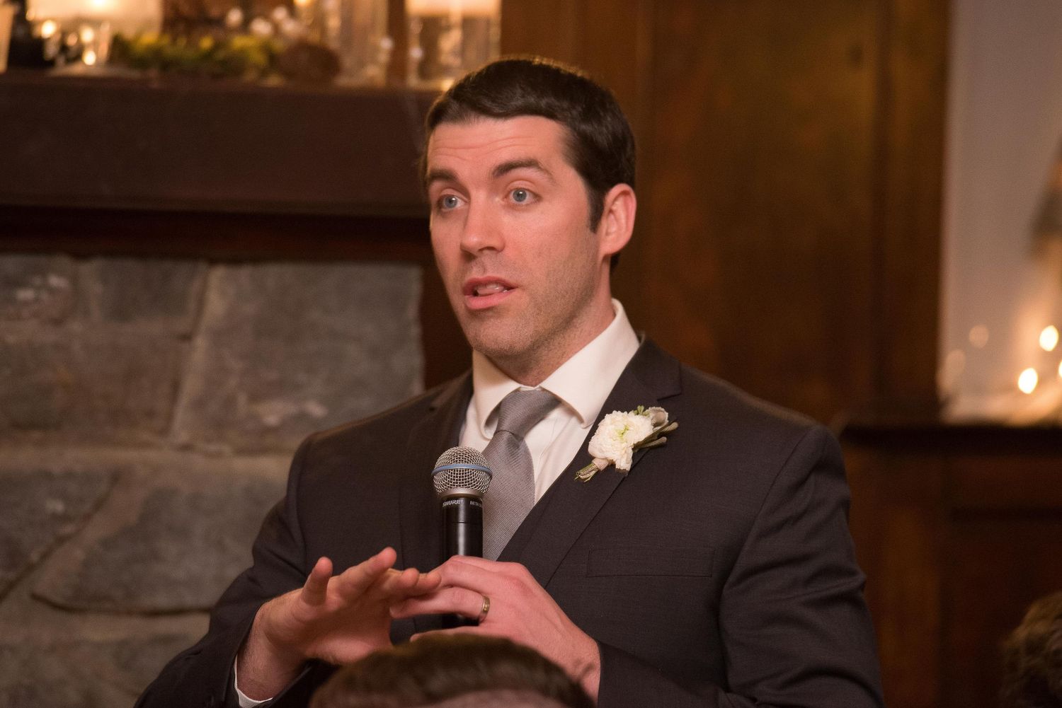 Man in dark suit with boutonniere speaks into microphone at wedding reception near stone fireplace.