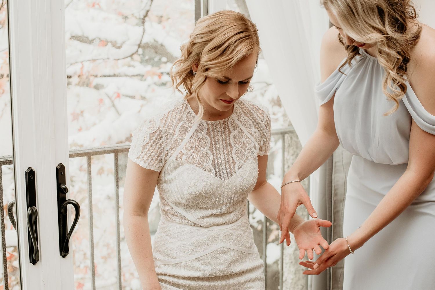 Bridesmaid in gray dress helping bride in lace wedding gown with bracelet near French doors.